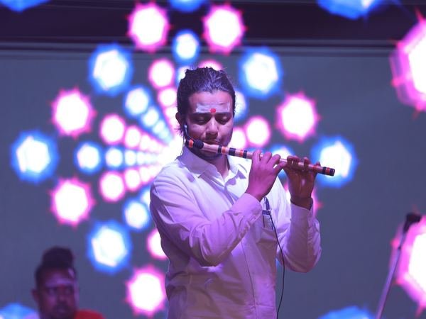 A flautist playing the bansuri on stage, with coloured stage lights behind him, at a music programme at Amartarangini, Haridwar